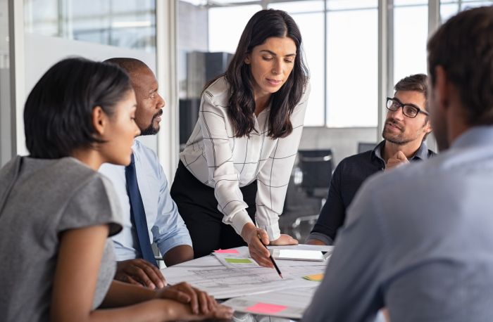Gruppe von Personen sitzt an einem Konferenztisch in einem modernen Büro und bespricht Dokumente. Eine Person steht und zeigt mit einem Stift auf ein Blatt Papier, während andere aufmerksam zuhören.